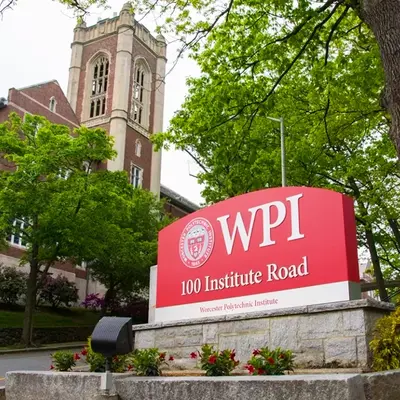 Eingangsschild des Worcester Polytechnic Institute (WPI) mit rotem Hintergrund und weißer Schrift, auf dem ‚WPI – 100 Institute Road‘ steht. Im Hintergrund ist ein historisches Universitätsgebäude aus rotem Ziegelstein mit einem Turm und gotischen Fenstern zu sehen, umgeben von grünen Bäumen und Pflanzen.