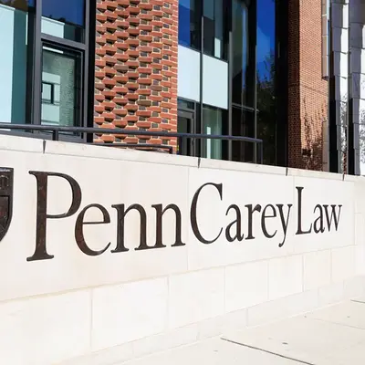 Außenansicht der University of Pennsylvania Carey Law School – Ein modernes Gebäude mit einer Fassade aus roten Ziegeln und großen Fenstern. Im Vordergrund ist ein helles Steinmauerstück mit dem eingravierten Schriftzug „Penn Carey Law“ und dem Wappen der Universität zu sehen.