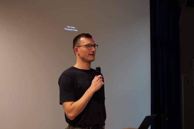 Viktor Hübner spricht mit einem Mikrofon in der Hand vor einer leeren Projektionsfläche.