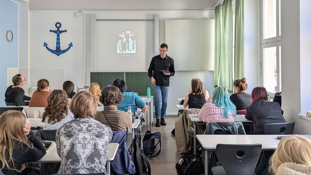 Viktor Hübner hält vor einer Schulklasse einen Vortrag. Die Schülerinnen und Schüler sitzen an Tischen und blicken zur Tafel, auf der ein Bild projiziert wird.